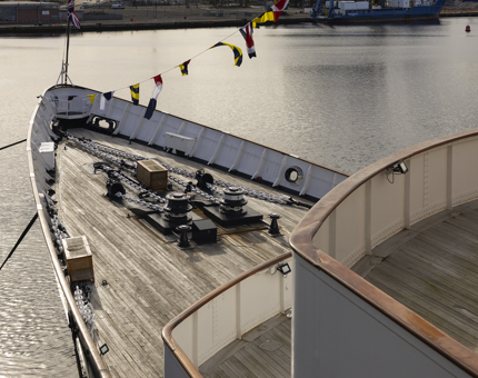 A view of Britannia's Bow from above in the evening sun. There are colourful dress flags hanging above. You can see the water of the Port of Leith in the background. 