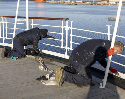 Two Maintenance men are kneeling on deck, painting the railings. 