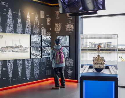 A visitor is in the Britannia's Visitor Centre looking at pictures of the ship being built. There is a model of Britannia in the foreground. 