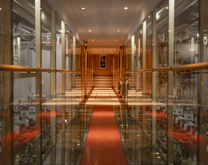 A corridor with reflective glass walls, showing a view of the ships Engine Room. The floor has a red walkway. 