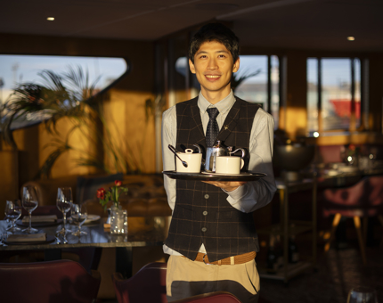 A Hospitality Server  in The Lighthouse Restaurant aboard Fingal is holding a tray with a silver teapot, cup and saucer and a sugar pot on it. 