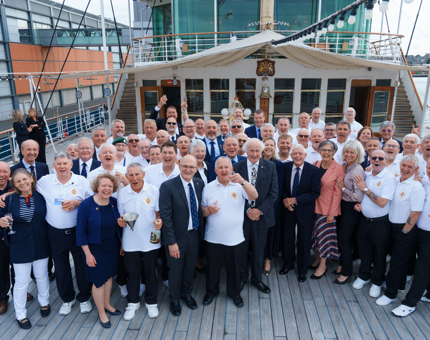 A large group of people including Royal Yachtsmen standing on the Verandah Deck outside the Sun Lounge. 
