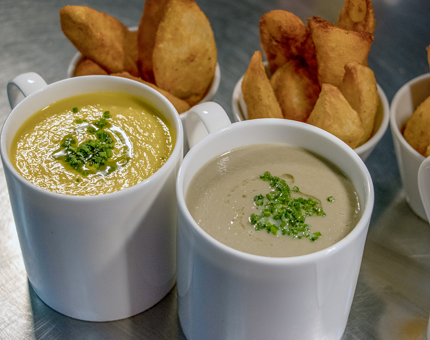 Two mugs of soup with garnish next to bowls of wedges.