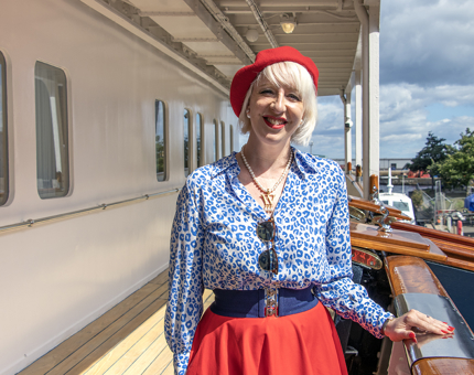 A woman wearing a blue blouse, red skirt and red hat is smiling next to a handrail on The Royal Yacht Britannia's Verandah Deck next to the Royal Brow.