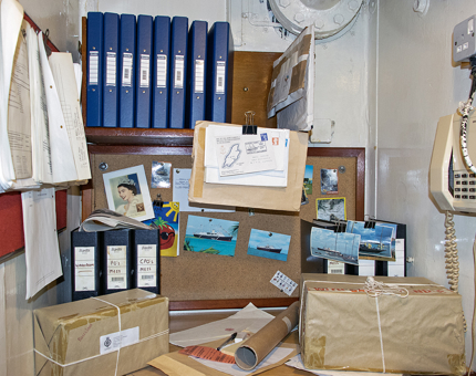 Inside Britannia's own Post Office. There are parcels on the table, postcards of the ship on a corkboard, a shelf containing ring binders and a phone on the wall.