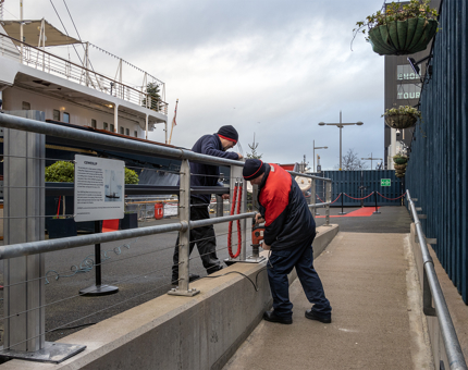 Two men from the Facilities team fixing a metal barrier to the concrete on the Quayside. 