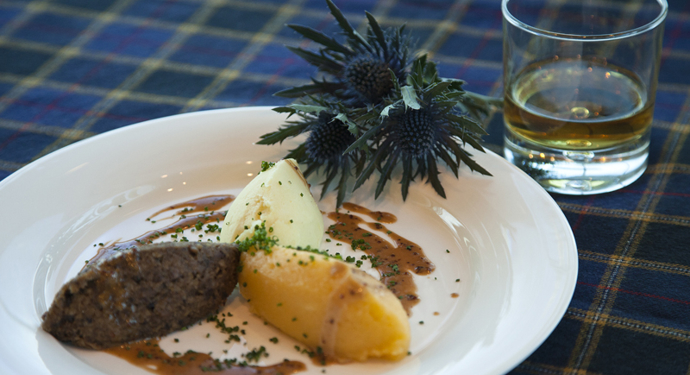 A plate of haggis, neeps and tatties and a glass of whisky on a tartan tablecloth.