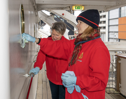 Two Housekeepers polishing a brass porthole and a handrail on the Blue Level of the Britannia tour route.