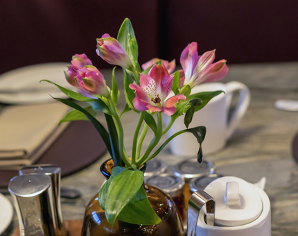 Pink flowers in a small brown glass vase are on a table in Fingal's Lighthouse Restaurant & Bar with place settings and cutlery. 