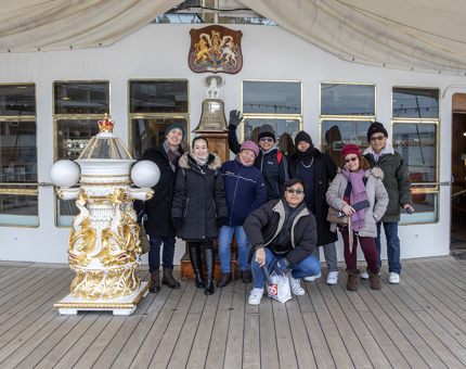 A group of visitors stand smiling on Britannia's Verandah Deck next to the Bell.