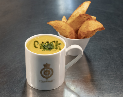A Britannia logoed mug containing soup and a small bowl of potato wedges next to it.