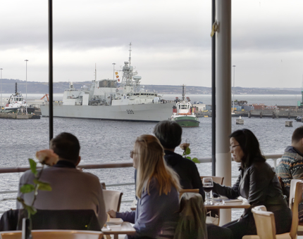 A view from inside the Royal Deck Tearoom. There are several visitors sitting at tables and a large floor to ceiling window with sea views and a large grey ship sailing past. 