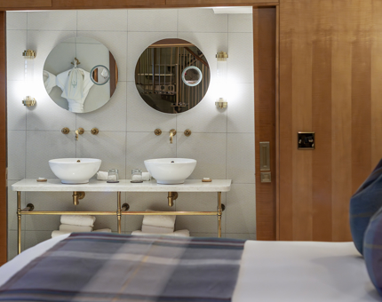 The bathroom in one of Fingal's luxury duplex cabins. There are two wash basins and mirrors. In the foreground is a bed with a blue tartan bed blanket and cushions. 