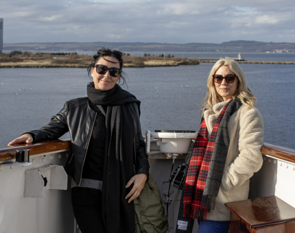 A woman wearing black and another woman wearing a red tartan scarf and sunglasses standing at the front of Britannia's Bridge smiling. 