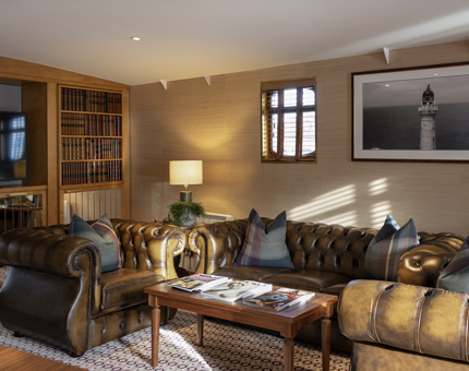 The inside of the Skerryvore Suite at Fingal, with brown Chesterfield sofa, armchairs, a coffee table, bookcase wall and a black and white photograph of a lighthouse on the wall. 