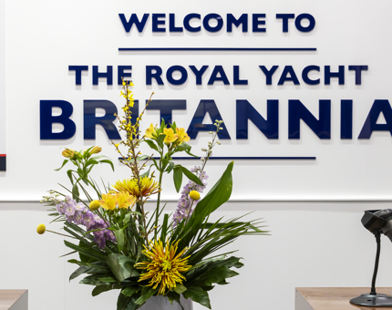 Signage for The Royal Yacht Britannia with a a large arrangement of flowers in the foreground.