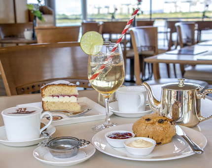A table in the Tearoom containing a silver teapot, cup and saucer, cocktail in a tall glass, slice of Victoria sandwich cake, fruit scone and a coffee with a Britannia ship stencil on top.
