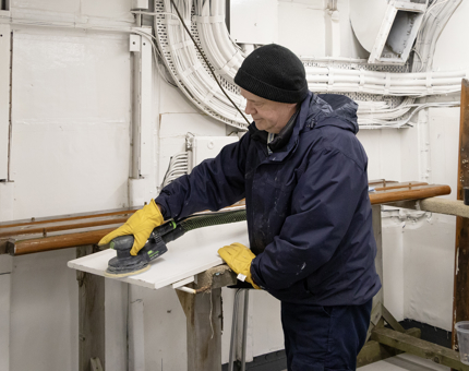 A member of the Maintenance Team uses a sanding machine on a door. 
