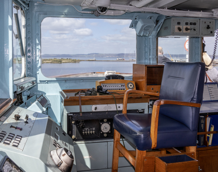 The Bridge on Britannia. There are dials and switches on a control panel, a tall leather chair, log books, charts and big windows.