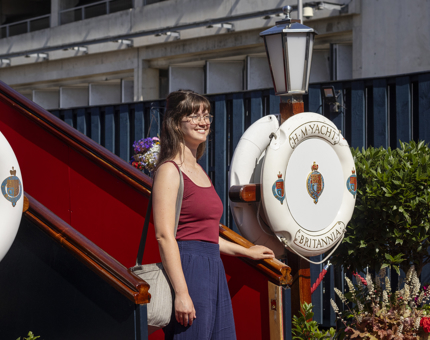 Visitor stands at the bottom of the steps to board the ship wearing a burgundy top and navy blue trousers.