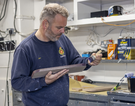 A man holding a paintbrush varnishing a piece of wood. 