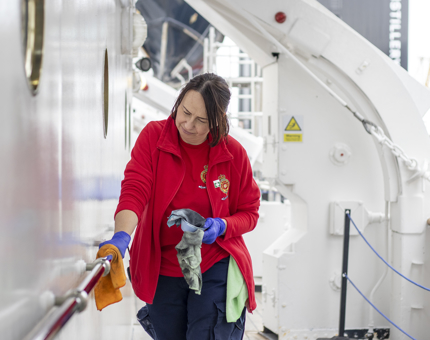 A woman in a red shirt and fleece polishing a silver handrail on the ships walkway.