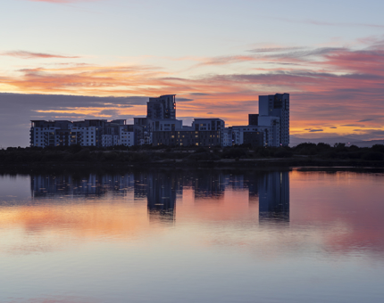 A view of the sunset over the water with a block of flats in the distance. 