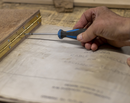 A hand holding a screw driver fitting a hinge to the ships drawings folder. 