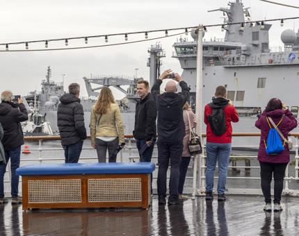 A groups of people standing on the Verandah Deck of The Royal Yacht Britannia watching a ship being towed our of the port. 