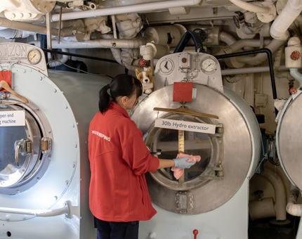 A woman Housekeeper holding a cloth cleaning a large washing machine in Britannia's laundry room.