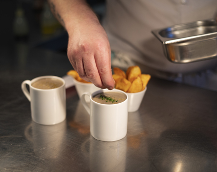 Two mugs of mushroom soup and two bowls of wedges. There is a chef placing cut chives on the soups. 
