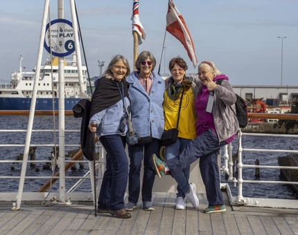 Visitors on the Verandah Deck posing for a photo. There is a ship in the background.