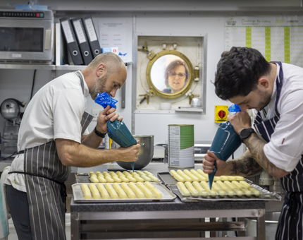 Two Chefs in the Galley preparing petits fours. A visitor looks on through the window in the background.