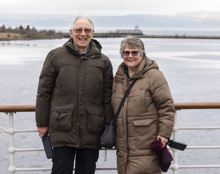 A man and a woman standing by the railings on the deck. There are views of the water in the background.