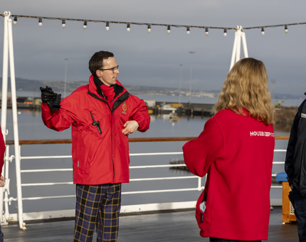 A man in a red jacket talks to a group of people. They are standing on the Verandah Deck. There is a view of the water behind them.