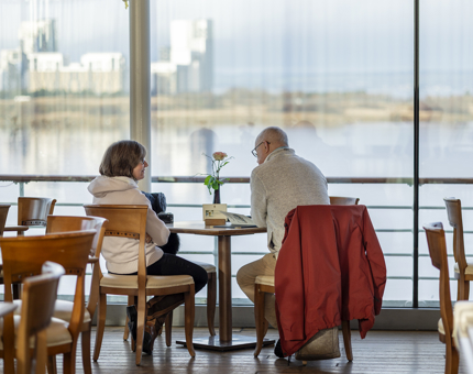 Two visitors sit at a table in the Royal Deck Tearoom. They are sitting facing large windows with a view of the waterfront.