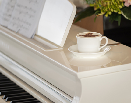A cappuccino with a chocolate stencil of a ship. It is sitting on a white grand piano. 