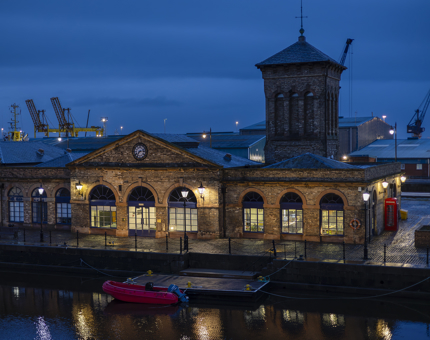 A view from Fingal Hotels deck. There is a brick building on the other side of the dock, it's lights are on, it is dusk and the sun has set.