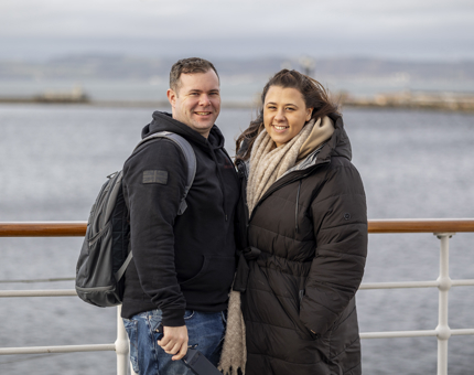 A man and a woman posing for a photo on the Verandah Deck. There is views over the water behind them.