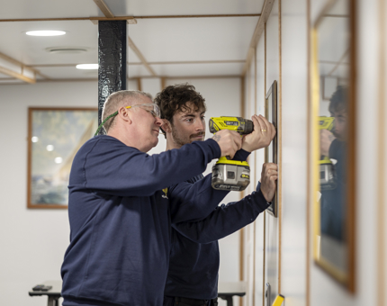 Two Facilities Officers hanging picture frames. On is holding a drill, the other is keeping the frame in place. 