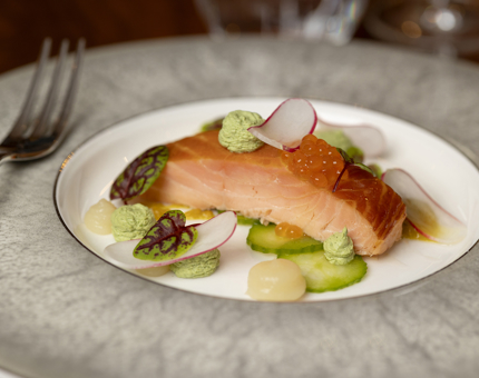 A grey and white plate with a piece of salmon on it. There is a fork sitting on the edge of the plate. 