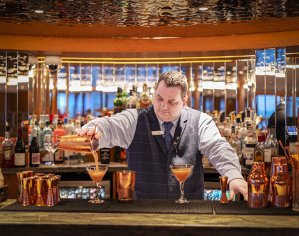 A man behind the bar aboard Fingal Hotel pouring a cocktail into a martini glass. The background of the bar is mirrored.