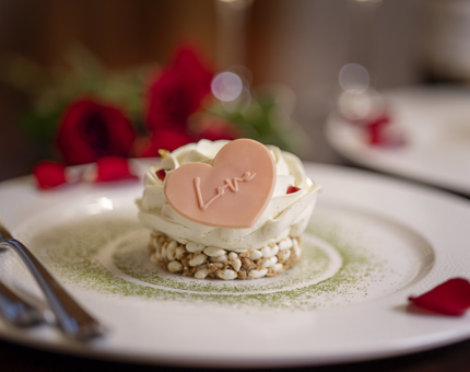 A white plate with a Valentine's dessert on it. There is a pink chocolate heart on it with the word love written on it. There are rose petals scattered on the plate and roses in the background.
