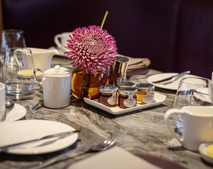 In Fingal Hotel's Lighthouse Restaurant & Bar, a table is set for breakfast. There is a tray with small pots of honey and jam, and a vase wit a large pink flower in it. 