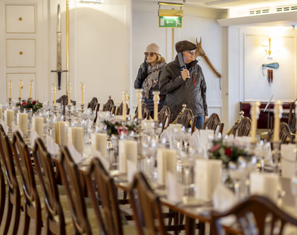 Two visitors are walking through the State Dining Room. They are holding the audio guide handset up to their ears. In the foreground is a long table with chairs, set for dinner. 