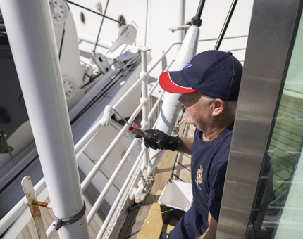 A Close up of a Maintenance man kneeling by a railing applying a coat of white paint. 