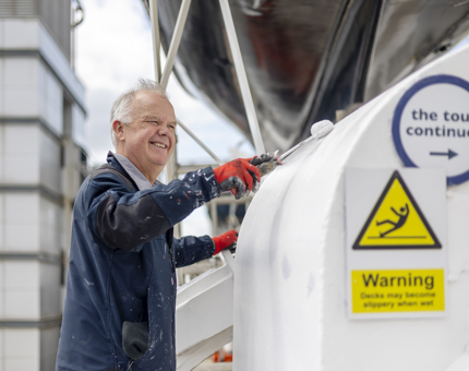 A maintenance team member holding a paint roller applying a coat of white paint to a part of the portside deck.