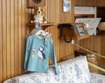 In Britannia's Sunlounge, a child's blue long sleeved top with a picture of a puffin on it is hanging from a shelf that has a plush corgi toy sitting on it. 