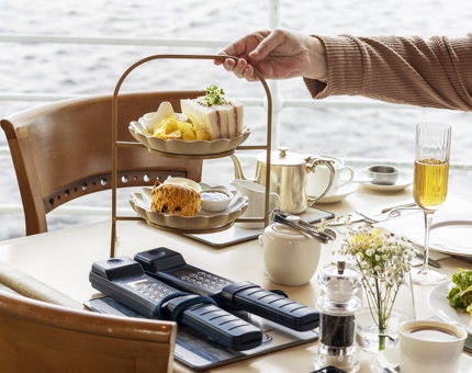 A hand is placing a cake stand with sandwiches and a scone onto a table in the Tearoom. 