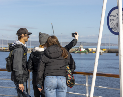 Three visitors on the Verandah Deck of The Royal Yacht Britannia taking a selfie. The Port of Leith is in the background. 